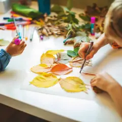 Niños de otoño creando arte con hojas secas y rotuladores.