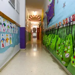Pasillo de guardería en Comunidad Valenciana. Dibujos infantiles decoran la pared, y mochilas verdes cuelgan listas para la ayuda.