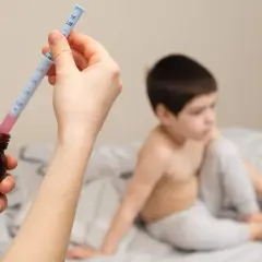 Mano preparando jeringa con ibuprofeno para un bebé. Niño en cama al fondo.