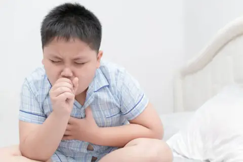 Un niño con pijama a cuadros tose con fuerza, agarrándose el pecho. Mi hijo no para de toser, parece que necesita ayuda.
