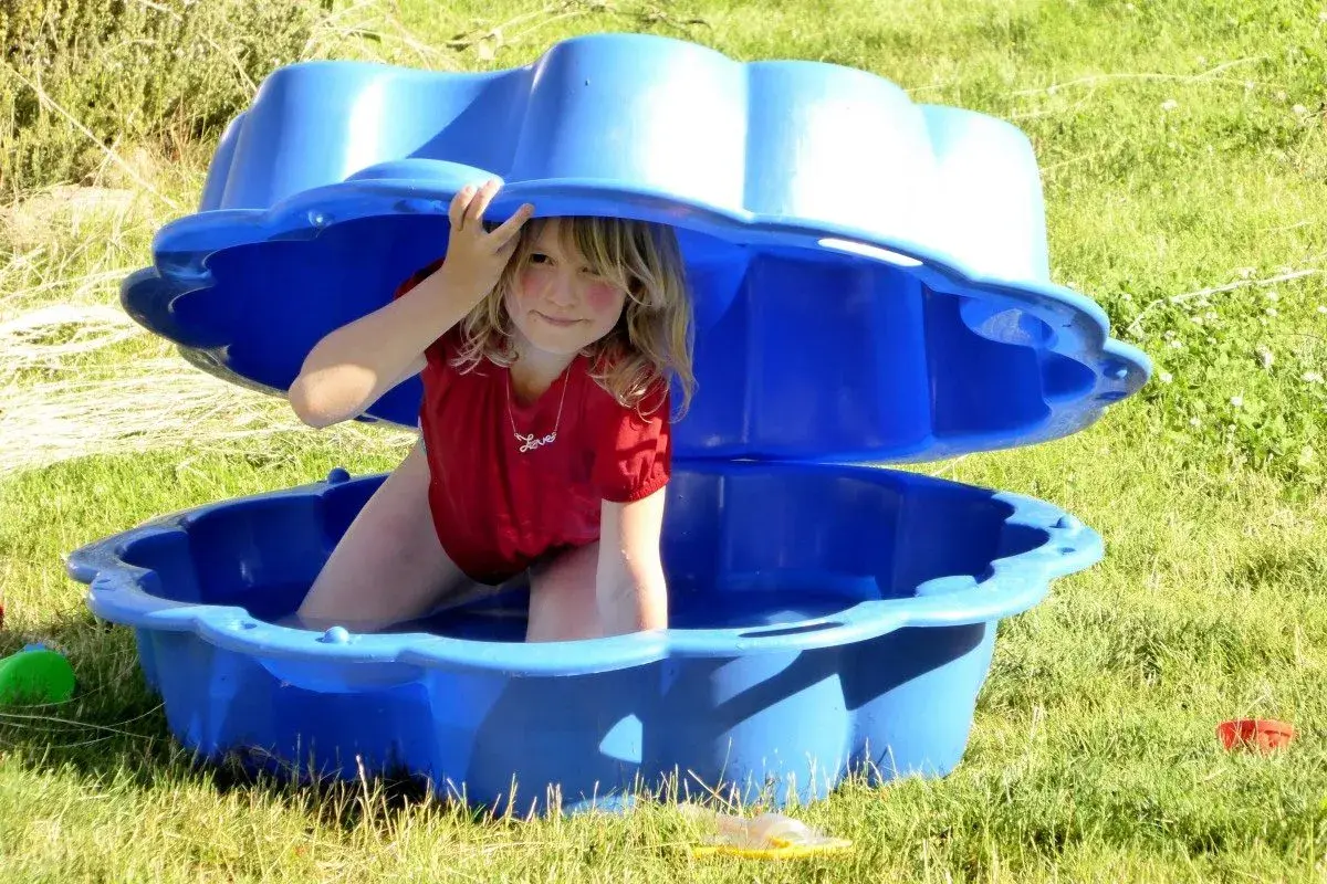Niña sonríe bajo una tapa azul de arenero, disfrutando de actividades de verano. ¡Diversión acuática para niños de 2 años!