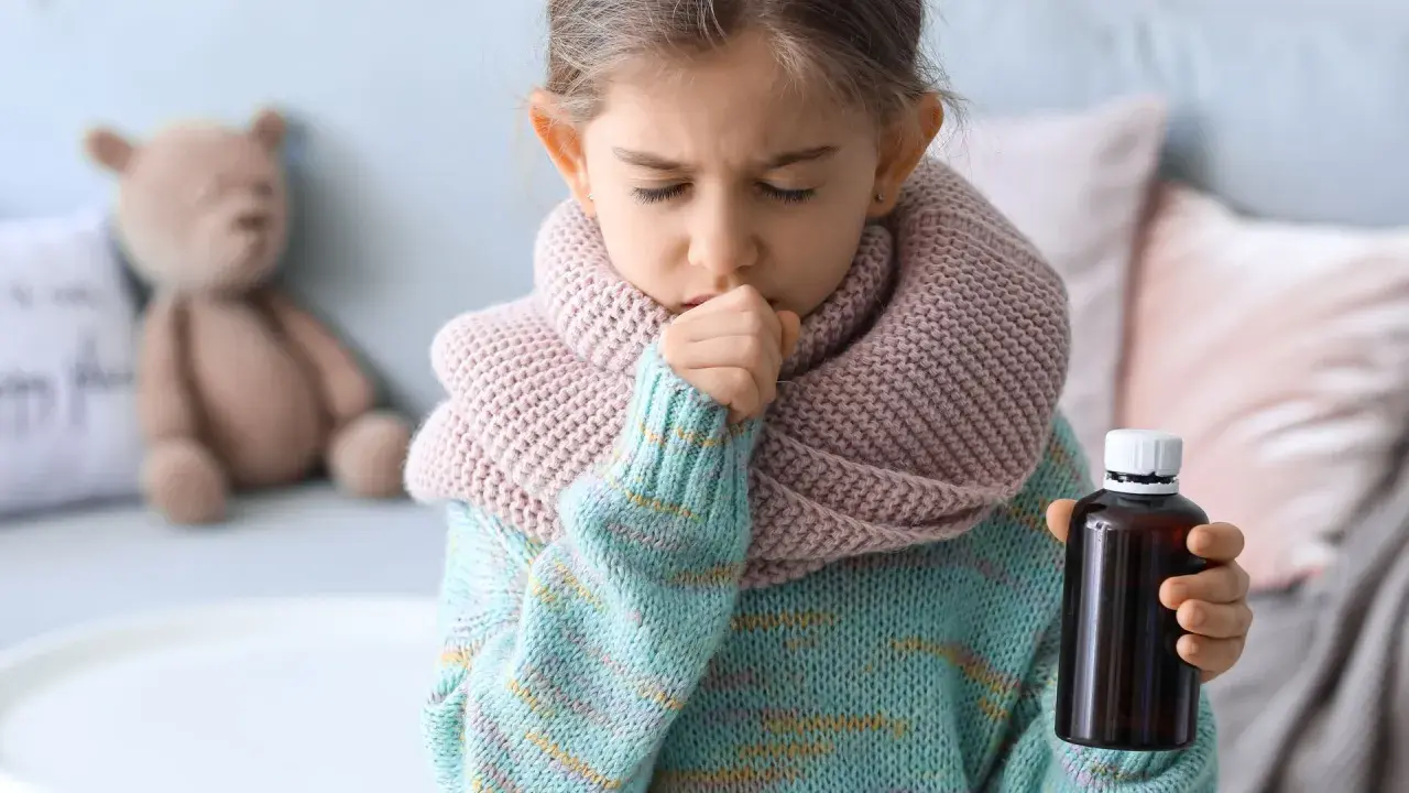 Niña de 3 a 4 años con tos, envuelta en bufanda y suéter, sosteniendo un frasco de medicina.
