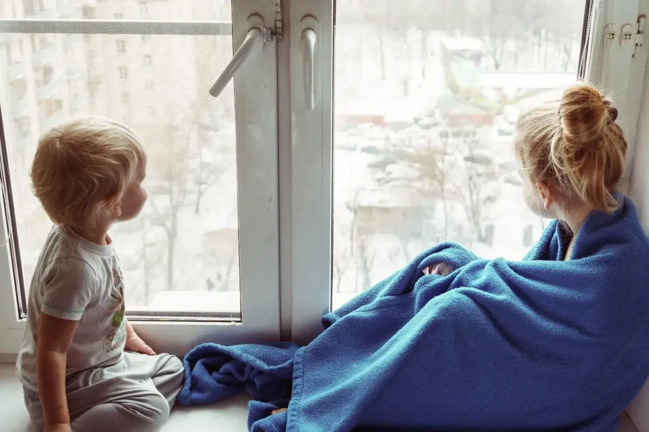 Dos ni&ntilde;os, uno de 3-4 a&ntilde;os, observan la nieve desde la ventana. Uno est&aacute; envuelto en una manta azul.
