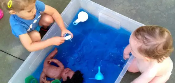 Niños de 3 a 4 años disfrutan de juegos acuáticos en el colegio, explorando con una muñeca y recipientes.